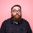 Man with glasses and a thick beard wearing a denim jacket against a pink background.